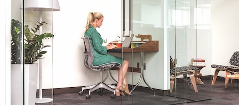 A person in a green dress is sitting in a Setu Chair and typing on her laptop at a small desk in a glass-walled workspace. Spaces like this give people the opportunity to foucs on heads-down work in an open office setting.