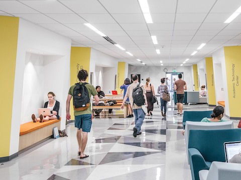 A corridor in an academic building; some students walk between classes while others relax in blue and gray Swoop Arm Chairs. 