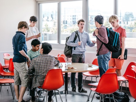 Seven male college students interact between classes, some sitting in red Eames Molded Fiberglass Chairs. Seven male college students interact between classes, some sitting in red Eames Molded Fiberglass Chairs.
