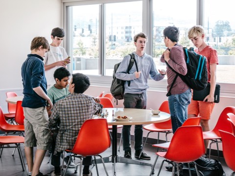 Seven male college students interact between classes, some sitting in red Eames Molded Fiberglass Chairs.