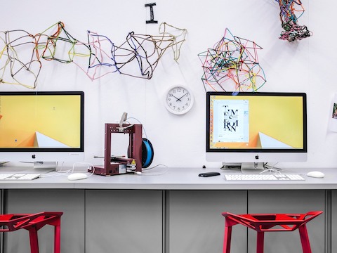 Abstract art decorates the wall behind two student workpoints outfitted with monitors, a shared work surface, and red stools. 