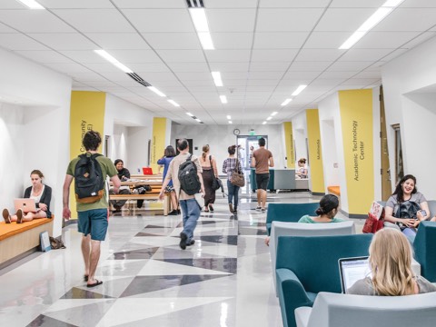 A corridor in an academic building; some students walk between classes while others relax in blue and gray Swoop Arm Chairs. 