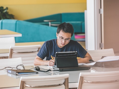 A college student works on a tablet computer while seated at a table serviced by white Eames Aluminum Group Chairs. 