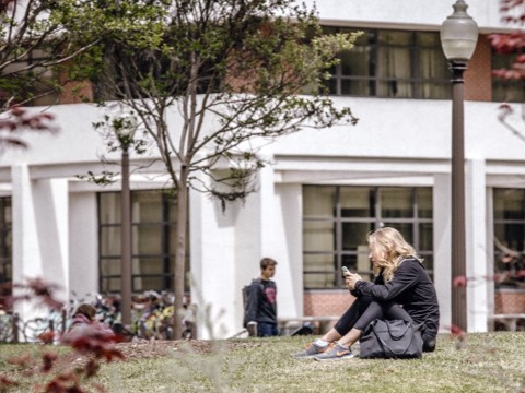 A female college student checks her smartphone while sitting on the grass outside an academic building. A female college student checks her smartphone while sitting on the grass outside an academic building.