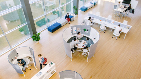 Four partially enclosed round gray Prospect spaces sit in a bright open office space, with workers collaborating or reading inside each space. A row of desks sit nearby with six white Sayl Chairs and blue and gray benches line the wall of windows.