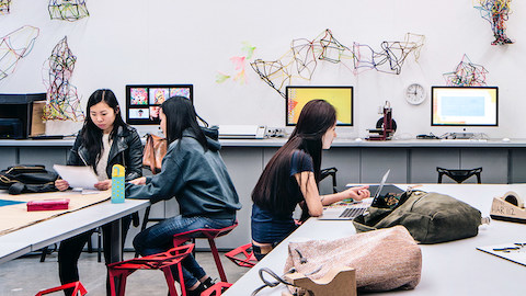 Three female college students sit atop red Magis Stool_Ones at tall desktops littered with backpacks and papers. A row of computer screens on tall destops sit against the back wall with 3D artwork displayed.