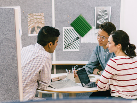 Espace collaboratif Prospect en demi-lune avec tissu punaisable gris et trois collaborateurs discutant autour une grande table ronde sur laquelle se trouvent un ordinateur portable, un magazine et des outils de dessin.