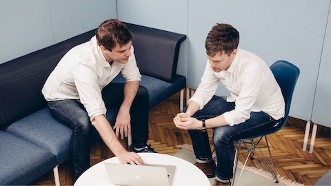 Two men in blue jeans and blue seating huddle over a laptop. Select to learn about the Living Office. 