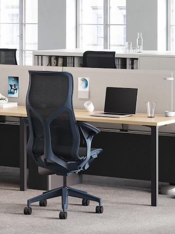 A dark blue Cosm high-back chair at a Canvas workstation with a graphite base, ash surface, and a tackable screen in gray. A dark blue Cosm high-back chair at a Canvas workstation with a graphite base, ash surface, and a tackable screen in gray.
