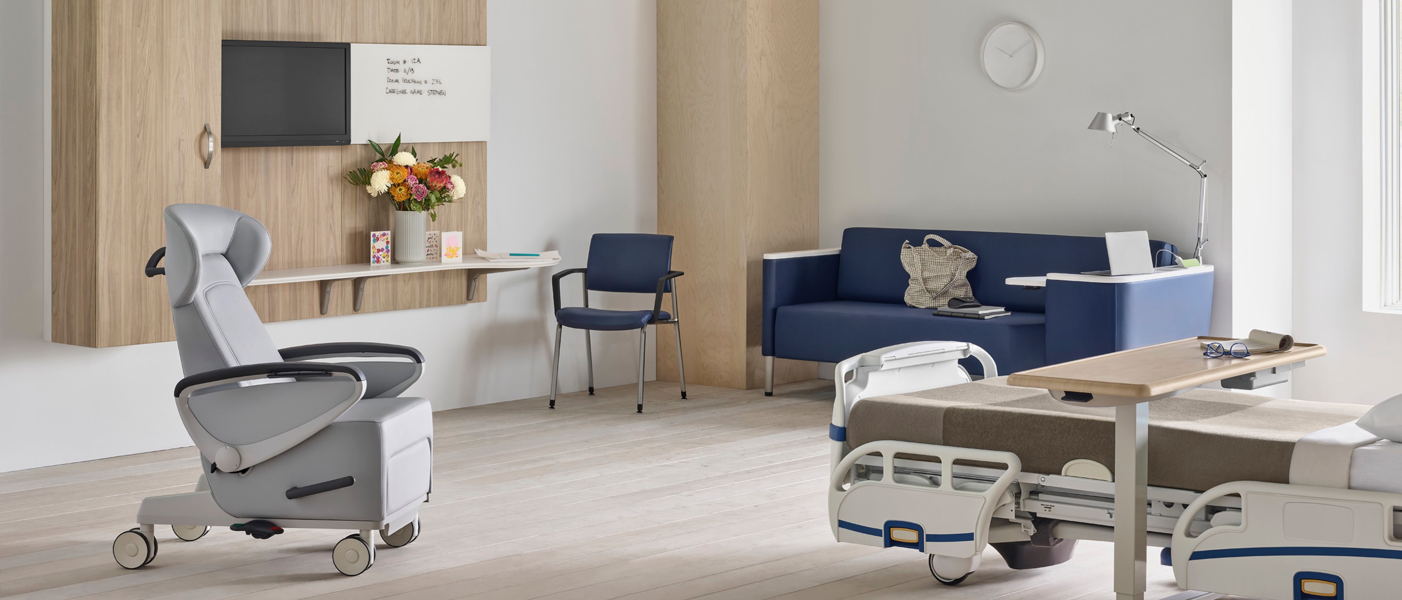 A Compass System footwall in a patient room with a gray Ava patient recliner, a blue Palisade Flop Sofa, a fully-upholstered blue Verus Side Chair, and a Mirage Overbed Table over a hospital bed.