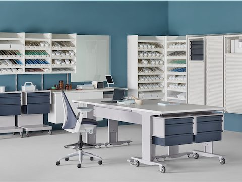 A pharmacy setting with a white Co/Struc System containing dispensing shelves, lockers, process tables, L Carts with blue drawers, and a Sayl Stool.