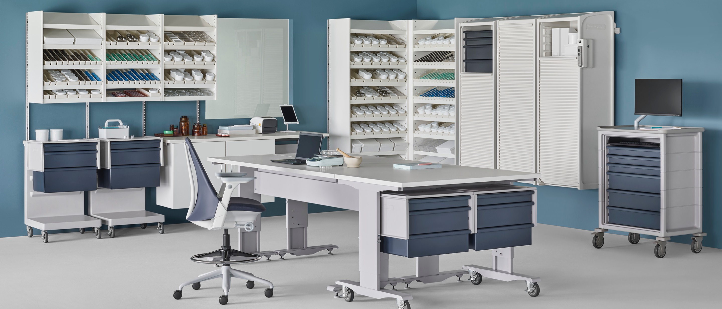 A pharmacy environment containing a Co/Struc System of dispensing shelves, lockers, process tables, L Carts with blue drawers, a technology cart with blue drawers, and a Sayl Stool.