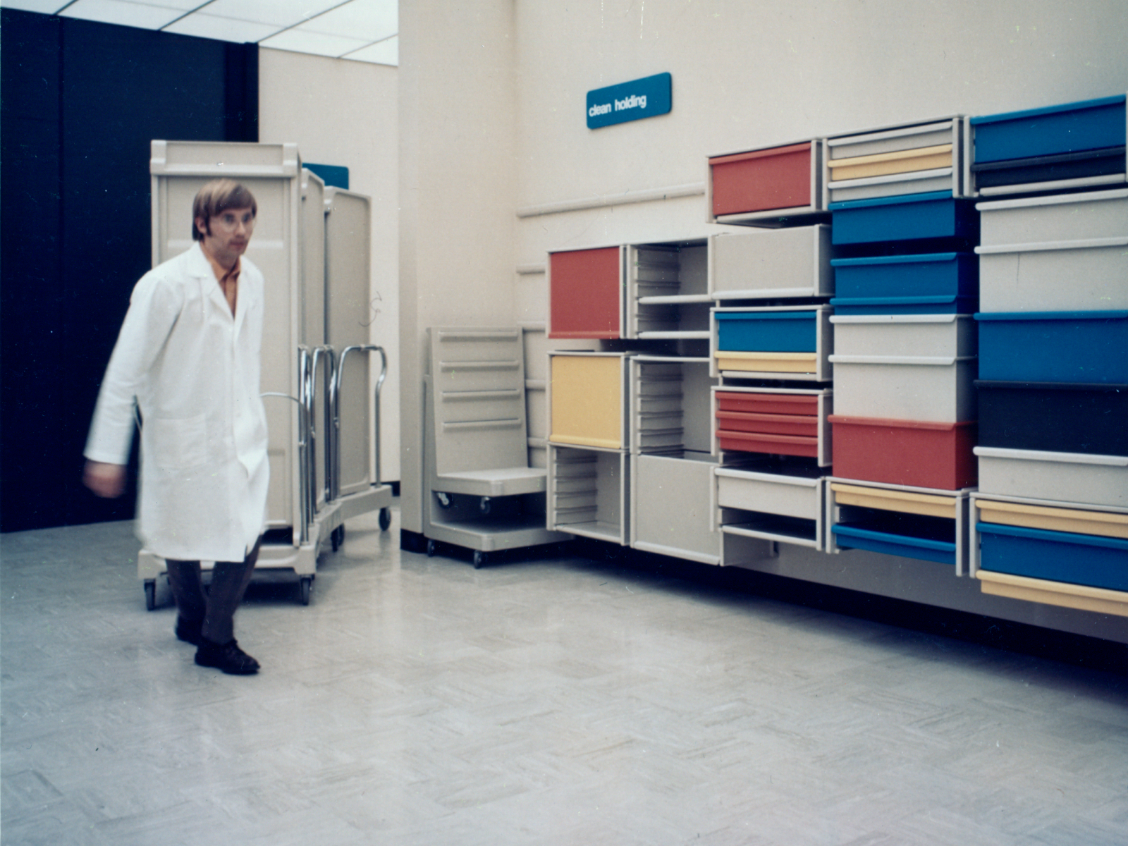 Archival photo of a man in white lab coat pulling Co/Struc lockers on transport cart in motion. Co/Struc wall-mounted open lockers with drawers in red, yellow, blue, and white background. Blue sign on the wall with white lettering that says 'clean holding.'