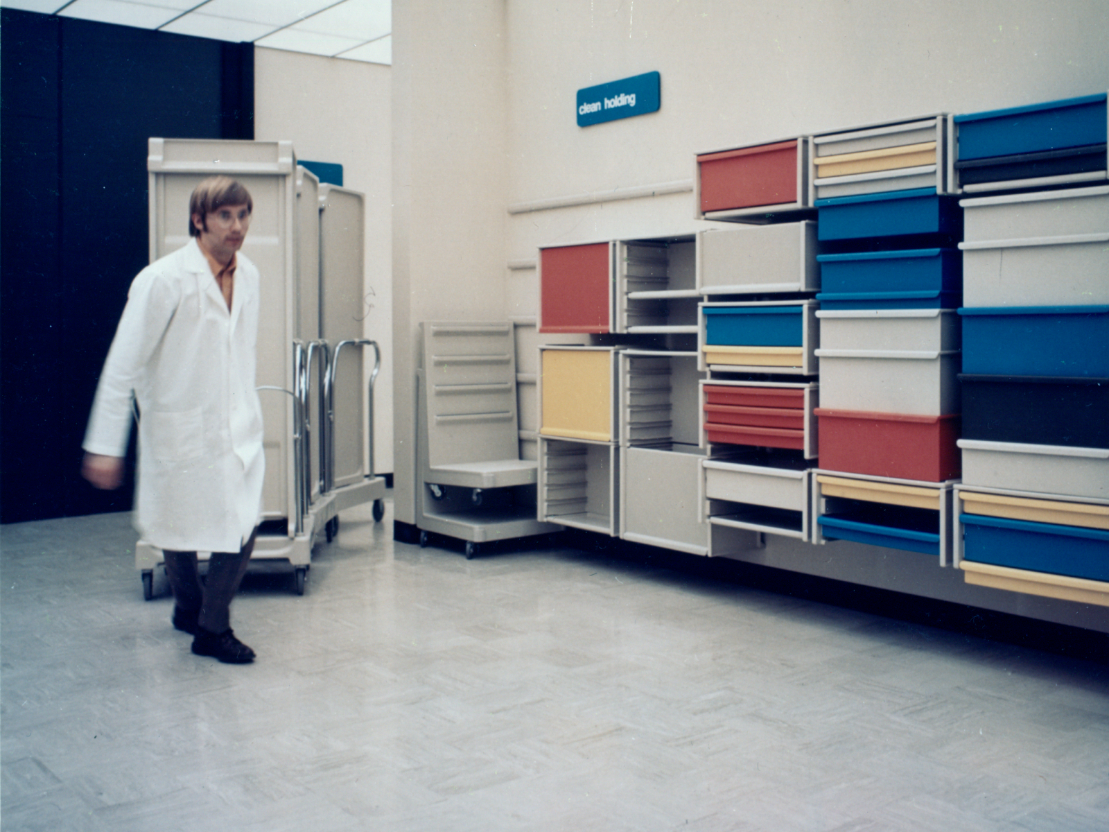 Archival photo of a man in white lab coat pulling Co/Struc lockers on transport cart in motion. Co/Struc wall-mounted open lockers with drawers in red, yellow, blue, and white background. Blue sign on the wall with white lettering that says 'clean holding.' Archival photo of a man in white lab coat pulling Co/Struc lockers on transport cart in motion. Co/Struc wall-mounted open lockers with drawers in red, yellow, blue, and white background. Blue sign on the wall with white lettering that says 'clean holding.'