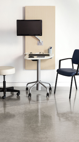 Exam room with Intent mobile table in a white top and silver base finish and wall unit in ash finish with a silver monitor arm holding a monitor. A Verus side chair in dark blue upholstery and a black frame. A physician's stool in a soft white upholstery and a black base.