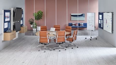 Seis Eames Soft Pad Chairs de color naranja quemado rodean una mesa en forma de lágrima en un área de colaboración con elementos de visualización Exclave. Seis Eames Soft Pad Chairs de color naranja quemado rodean una mesa en forma de lágrima en un área de colaboración con elementos de visualización Exclave.