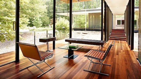 Een lichte zithoek met kamerhoge ramen, twee Sled-stoelen, een Tuxedo-bank en een I-tafel met glazen blad bijzettafel.