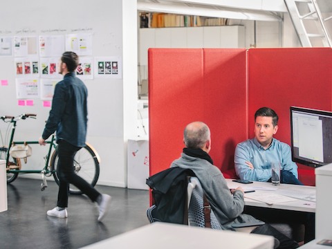 Two colleagues converse across a table in a workpoint bordered with a red privacy screen.