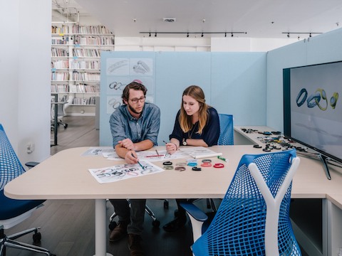 A man and woman discuss a project in a collaboration space featuring a peninsula table, video screen, and blue Sayl office chairs.