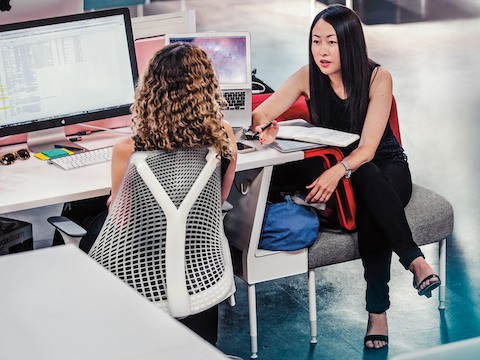 Two women co-workers converse while seated at a Public Office Landscape workstation.