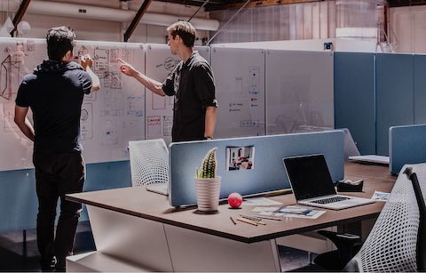Two men stand before a whiteboard, one drawing and one pointing. Two men stand before a whiteboard, one drawing and one pointing.