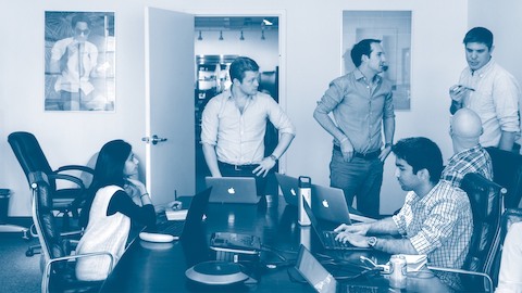 Six colleagues—three seated; three standing—interact in a conference room.