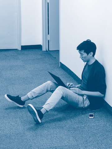 A man works on his laptop while seated on the floor of a hallway.