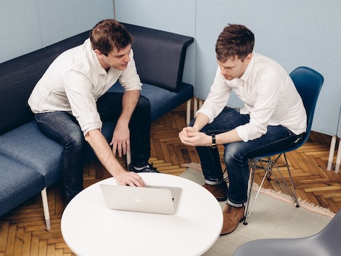 Two men view a laptop, one sitting on a blue Public Office Landscape sofa; the other on a blue Eames Molded Plastic Chair.