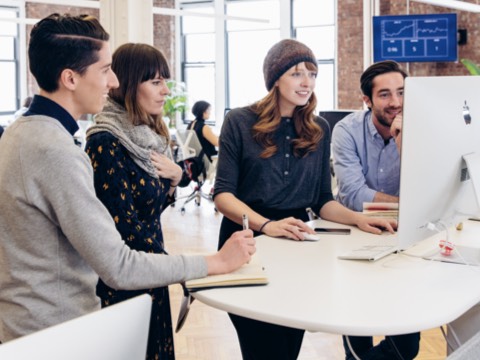 Two women and two men gather round a standing-height table to view a computer monitor. Two women and two men gather round a standing-height table to view a computer monitor.