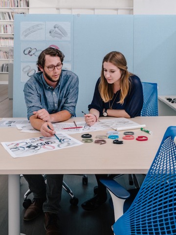 A man and woman sitting in blue Sayl office chairs lean on a table to discuss a project.