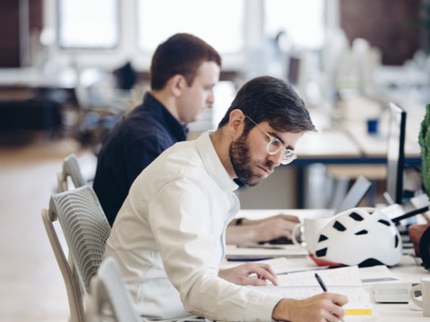 Two men in Sayl office chairs work next to each other in an open office with a benching setup. Two men in Sayl office chairs work next to each other in an open office with a benching setup.