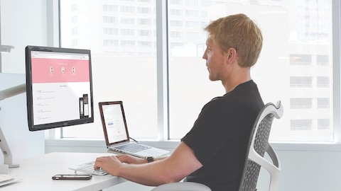 A casual office worker achieves correct ergonomic posture with a gray Mirra 2 chair and a Flo Monitor Arm.