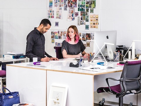 A standing man and seated woman collaborate at a shared work surface. A standing man and seated woman collaborate at a shared work surface.