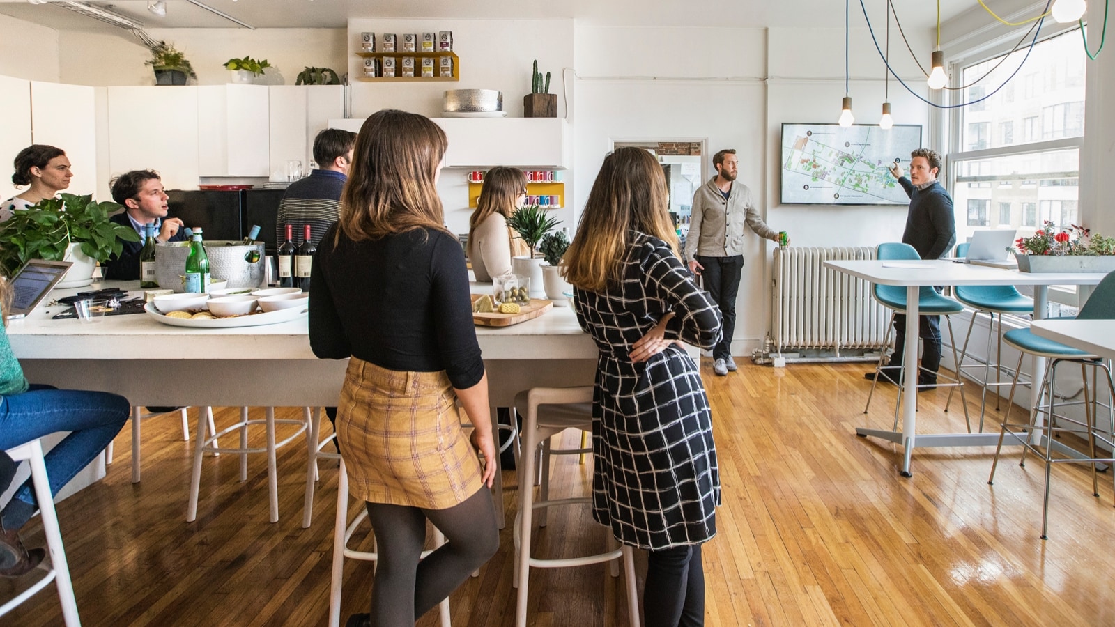A group of people listening to a person address them in an open office kitchen space.