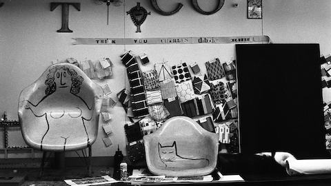 A black-and-white archival photo taken at Eames Studio of two fiberglass shell chairs with Steinberg's hand-painted designs, one of a nude reclining woman and the other a cat, in front of a wall of fabric swatches and a "Thank you Charles" sign.