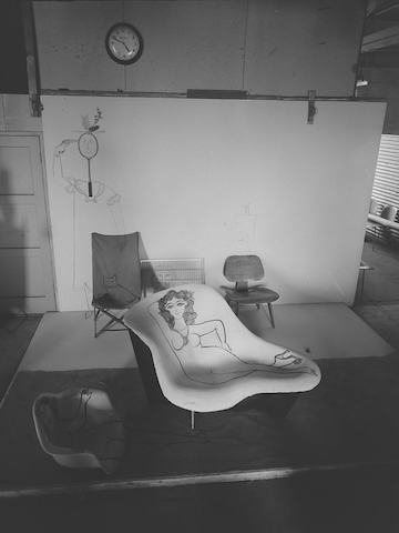 A black-and-white archival photo taken at Eames Studio of a curvy nude woman is hand-painted on a fiberglass lounge chair prototype, surrounded by a shell armchair, an LCW, and folding chair with various figures also painted upon them and extending to the white walls behind.