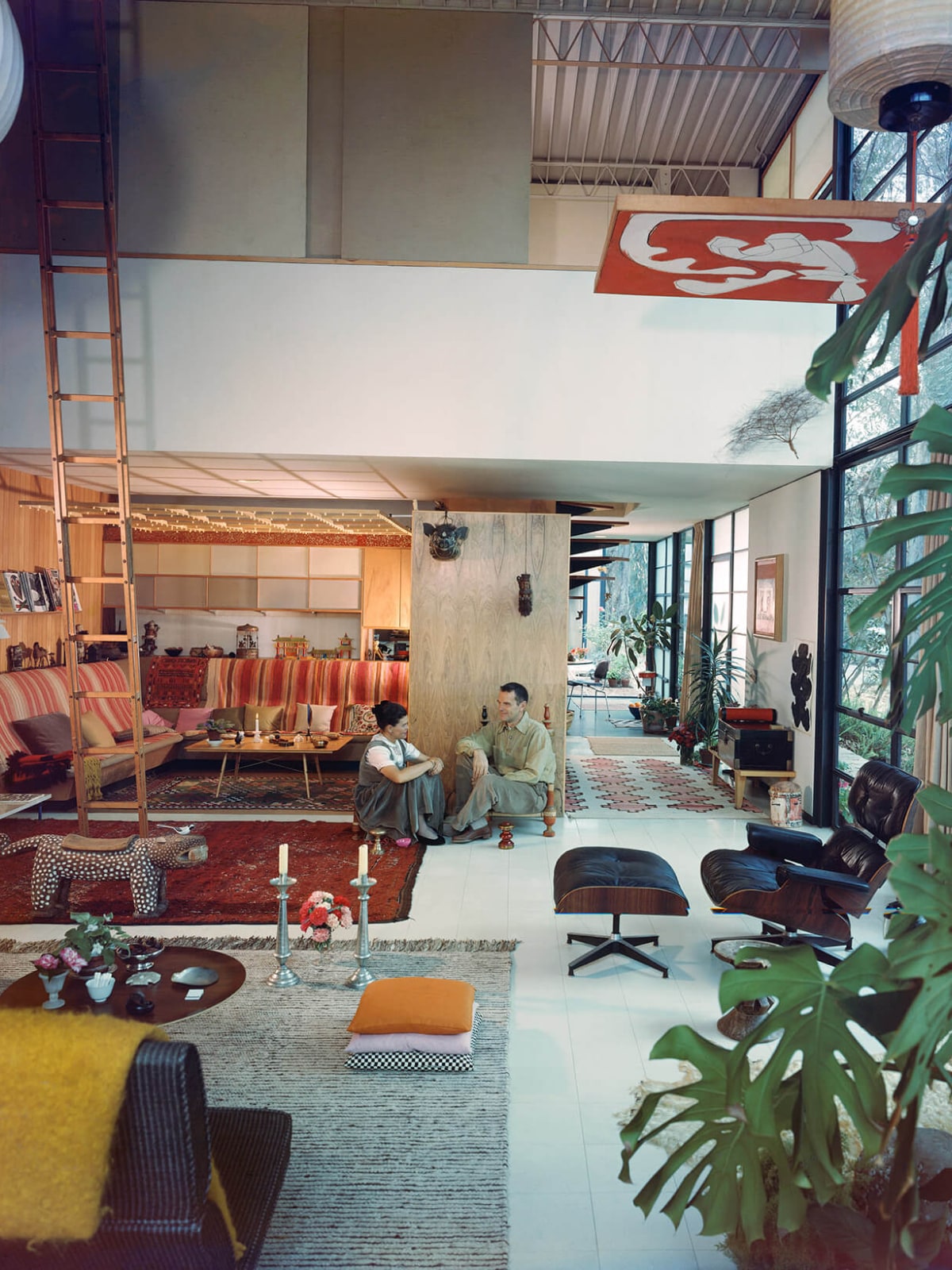 A colorful archival photo of Ray and Charles Eames sitting on the floor of their lofted ceiling living room, surrounded by candlesticks, objects collected from their travels, colorful pillows, and many of their pieces of furniture, including an Eames Lounge Chair and Ottoman in black leather.