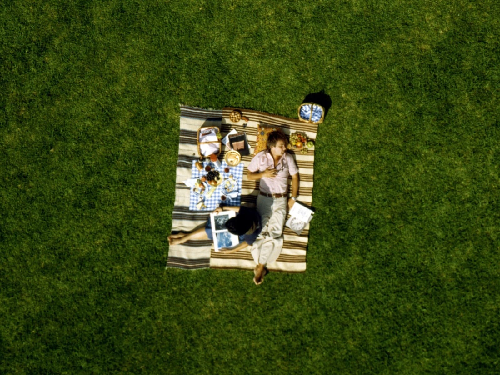 An overhead shot of a man and woman laying on a striped blanket with picnic food on green grass.
