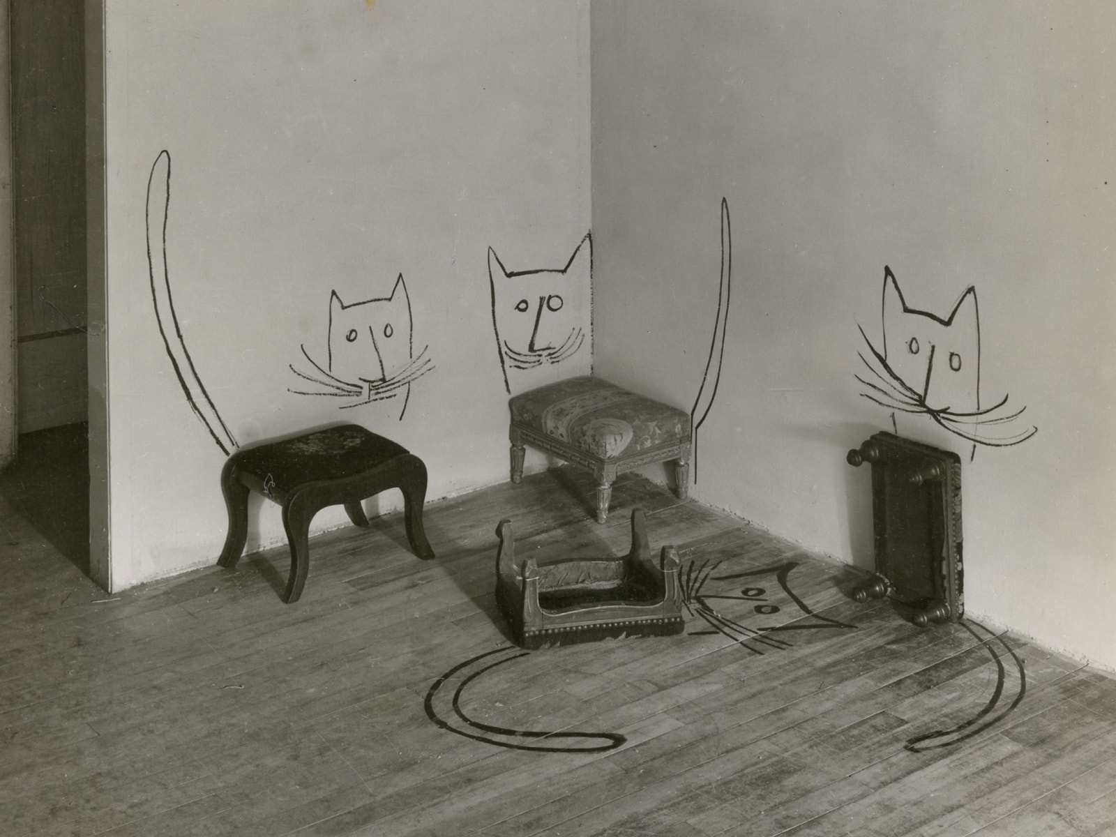 A black-and-white archival photo shows four footstools sitting in the corner of an empty room, with cats painted on the white walls and wood floors around them: two right side up, one upside down, and one set on its short side, feet facing away from the wall.