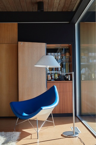 Sunshine pours in through a window of Craig Ellwood's Hunt House, bathing wood-paneled room featuring a blue Nelson Coconut Lounge Chair. 