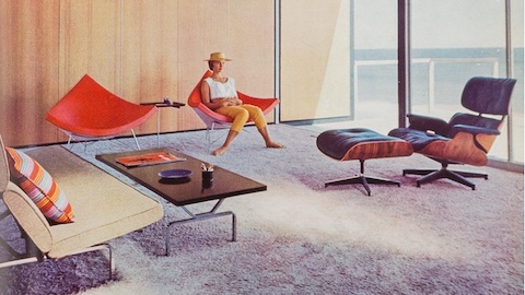 A woman sits in one of a pair of orange-pink Nelson Coconut Chairs in the sunny living room of a mid-century modern home in Malibu. An Eames Lounge Chair and Ottoman and Eames Sofa Compact are nearby.