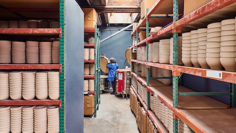 Stacks of unglazed bowls and plates sit on red and green shelving with a glazer in the background readying pieces.