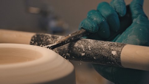 A close-up shot of a gloved hand holding a shaping tool against a piece of greenware, or dried-out clay, spinning on wheel.