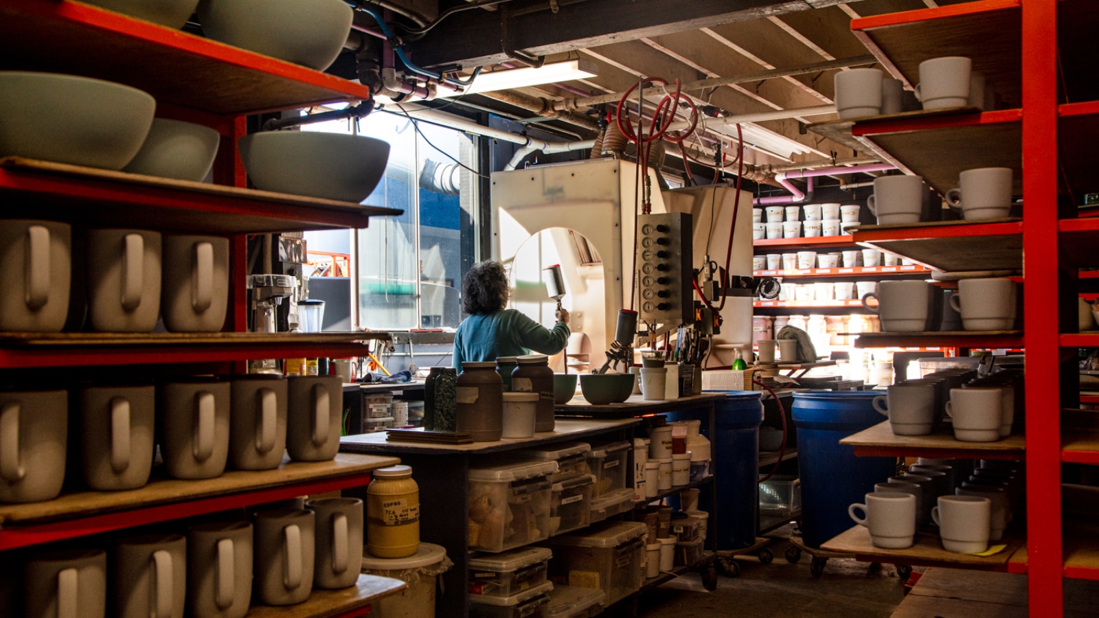 A view of many dried clay pieces including bowls, mugs, and plates, waiting on red shelves to be glazed and then fired.