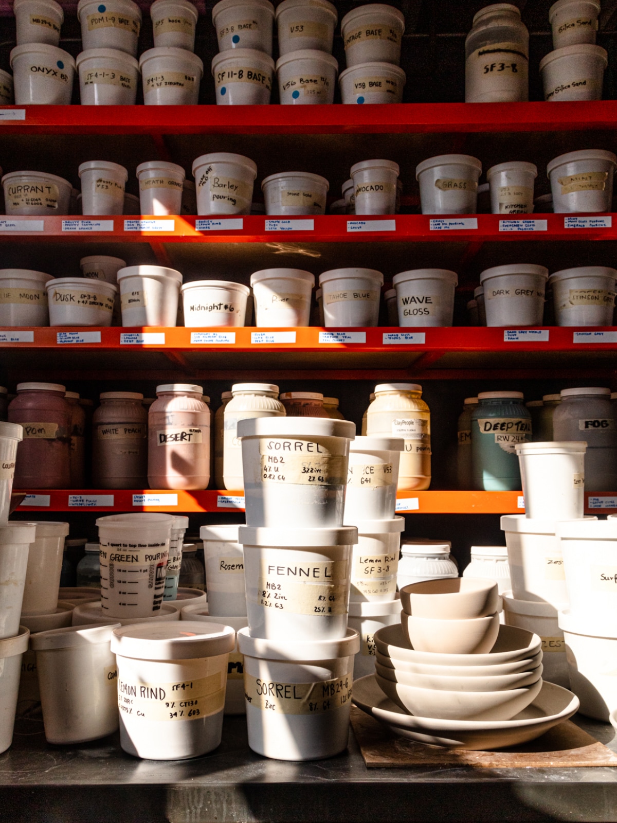 Red shelving with small white tubs featuring different labels with glaze coloros and types, with a stack of unglazed pottery next to it.