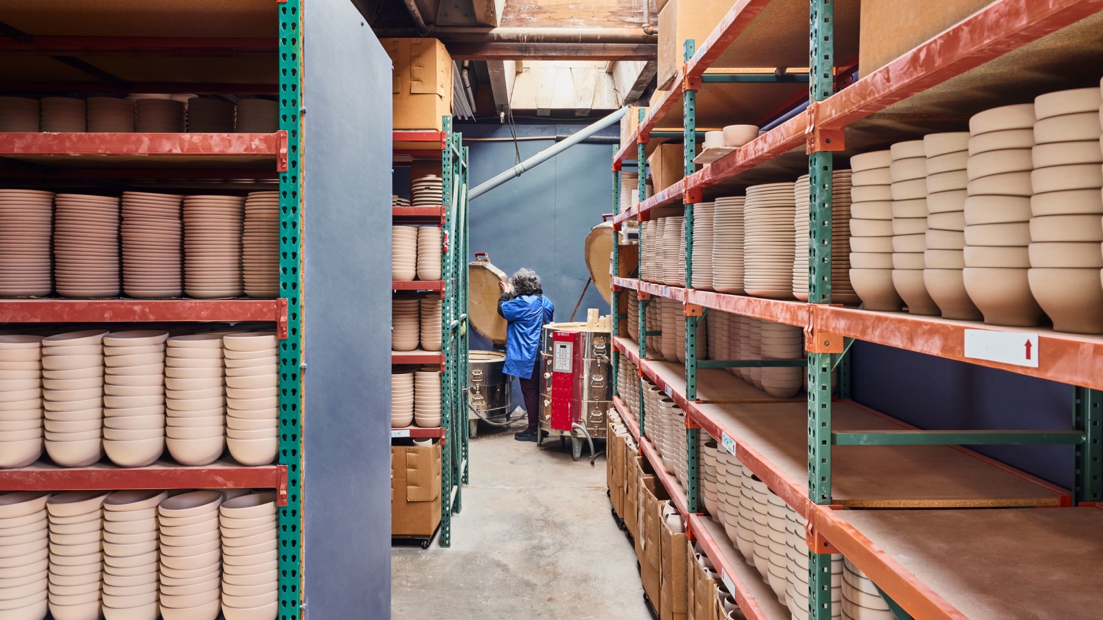 Stacks of unglazed bowls and plates sit on red and green shelving with a glazer in the background readying pieces.