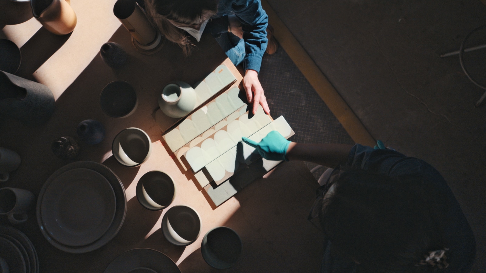 An overhead shot of two production team memberss pointing at reference pieces of pottery wtih various stoneware next to them on a table.