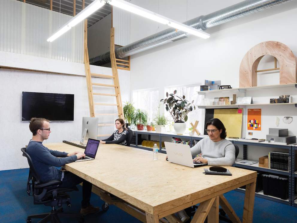 A woman and two men work on computers at a large table in a bright studio.