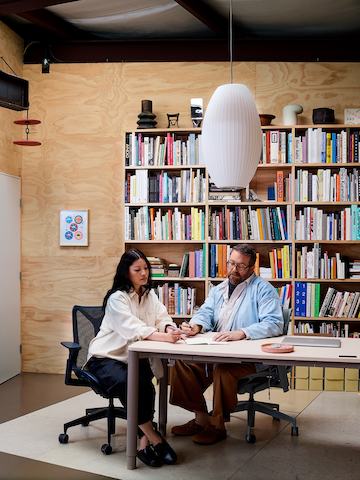 Heidi Korsavong and Benjamin Critton sit in dark gray Cosm office chairs at a white Spout Sit-to-Stand Table, with a full bookcase attached to plywood walls behind them and a Nelson Cigar Bubble Pendant Lamp above.