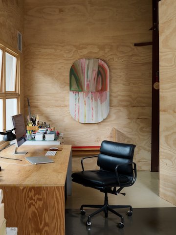 A black leather Eames Soft Pad Chair sits at a plywood desktop with computer and desk accessories, with modern art hanging behind on the plywood walls.